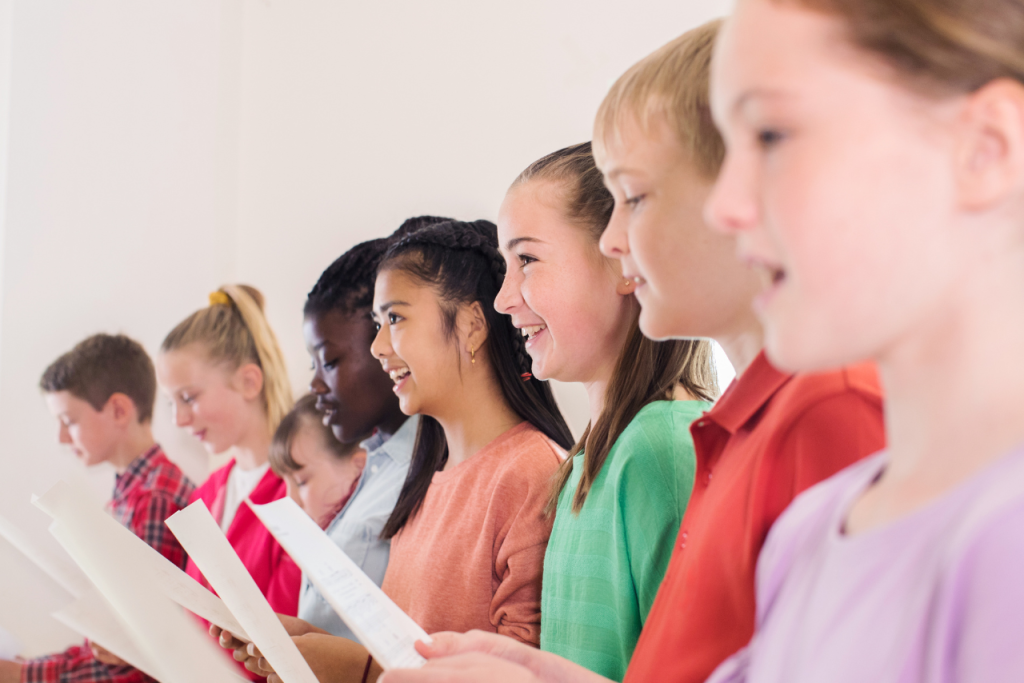 Group of smiling children and teens holding sheet music while singing together in a brightly lit classroom, representing joy, diversity, and teamwork in music education.